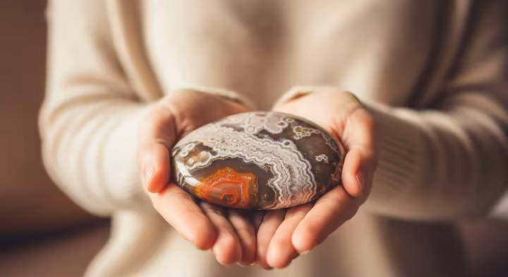 A person's hands gently cupping a polished Crazy Lace Agate palm stone, conveying a sense of calm and joy.