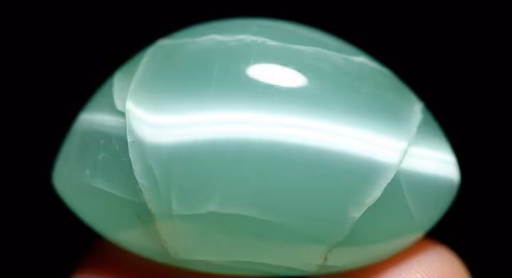 A polished cabochon of chatoyant green calcite showing a distinct cat's-eye effect across its surface.