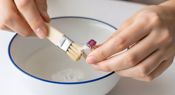 Hands gently cleaning a pink tourmaline ring with a soft brush and soapy water.